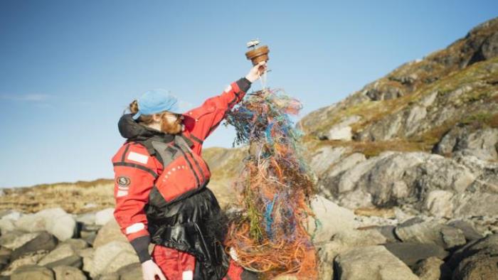 Vyras su gelėbjimosi liemene ir kompbinezonu laiko pakėlęs plastiko žuvims gaudyti tinklą į kurį įsipainiojęs siųstuvas